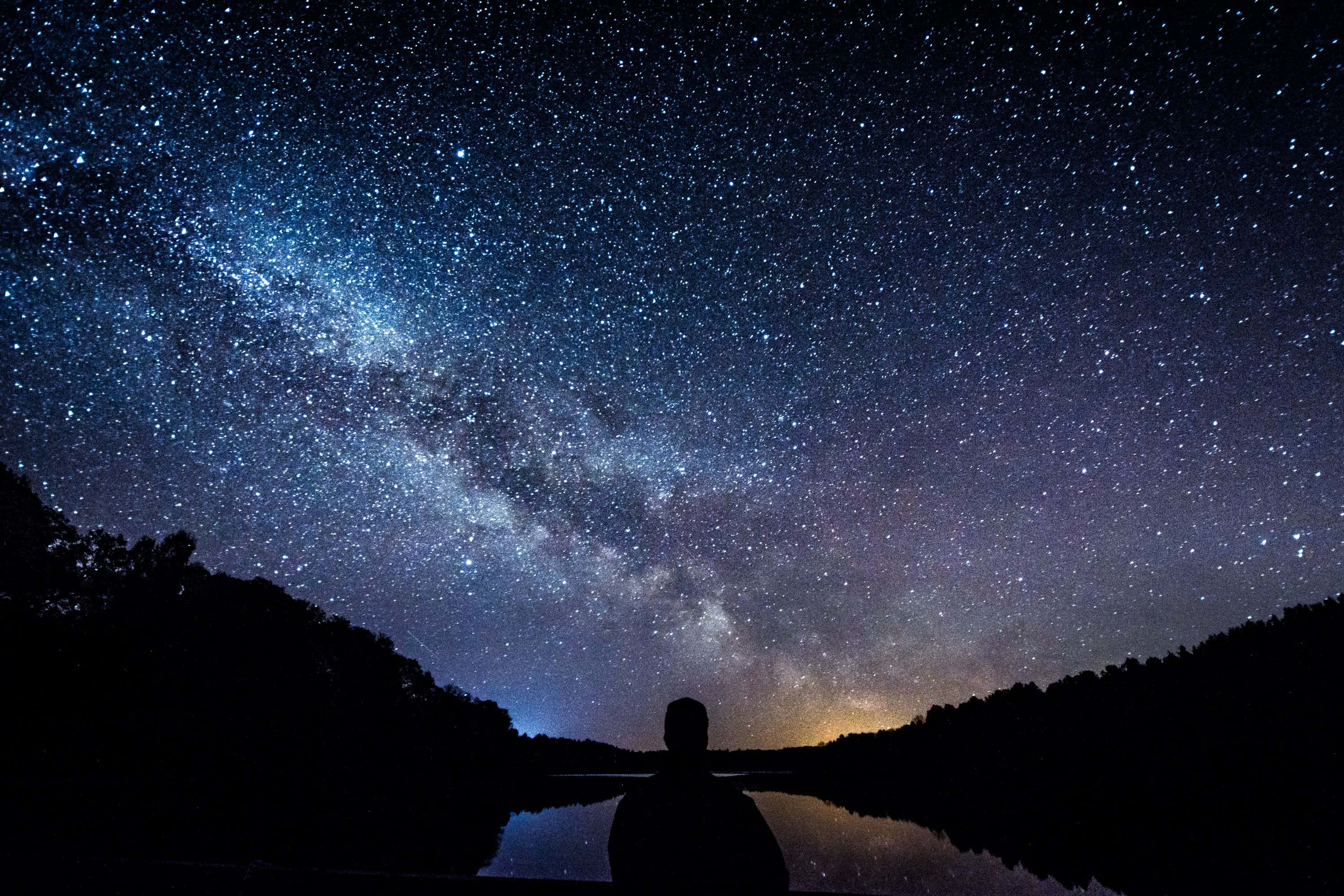 Person silhouetted against starry night sky above a calm lake surrounded by hills.